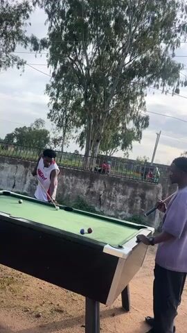 Two young men are playing billiards outdoors in Ban, Nigeria. The man on the left is wearing a white tank top with "HEAT" written on it in red and blue, and blue jeans. He is in the process of taking a shot with a pool cue. The man on the right is wearing a purple t-shirt and black pants. He is holding his cue and watching his opponent. The billiard table is green and appears to be in good condition. There are several billiard balls on the table, including a white cue ball, a red ball, and a blue ball.

The setting is outdoors, with a dirt ground and a concrete wall with a fence behind it. Tall trees with sparse foliage are visible in the background. The sky is cloudy, suggesting it might be overcast or late in the day. The overall atmosphere appears casual and recreational. The activity is a game of billiards, with one player actively playing and the other observing. The emotions seem neutral, focused on the game. The time of day is not explicitly indicated, but the lighting suggests it could be daytime, possibly late afternoon. The location is identified as Ban, Nigeria.