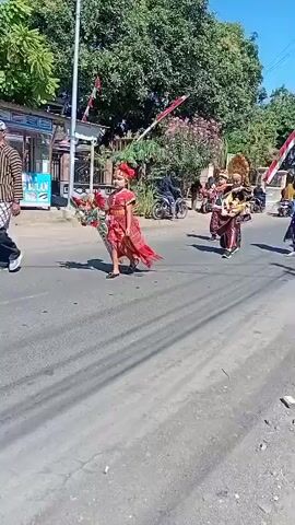 A child dressed in a red and gold traditional outfit walks down a street holding a bouquet. They are part of a parade, with other participants dressed in various costumes and carrying items like framed pictures and flags. The scene appears to be outdoors on a sunny day, with trees and buildings visible in the background.
