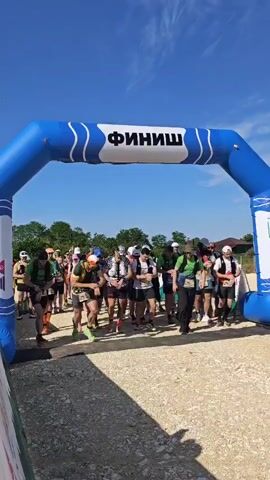 A group of approximately 30-40 runners, dressed in athletic wear and carrying hydration packs, are captured at the start of a race under a clear blue sky. They stand on a light brown dirt or gravel path, preparing to run through a large blue inflatable arch with "ФИНИШ" (FINISH) written in white Cyrillic letters across its top. Green trees and foliage are visible in the background, with a few distant buildings. The video begins with a countdown "3, 2, 1, START!" in Russian, after which the runners, wearing race bibs, begin to run forward, passing under the arch and towards the camera. Many of the participants are wearing sunglasses or hats.