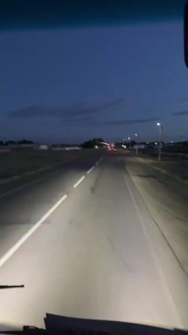 This video shows a point-of-view perspective of driving down a two-lane road at dusk or night. The road is paved and has white lines marking the lanes. The headlights of the vehicle illuminate the road ahead. On either side of the road, there are fields and some scattered buildings and utility poles. The sky is a deep blue with some wispy clouds. There are streetlights visible along the road, suggesting it may be near a town or settlement. The overall mood is calm and serene, with the headlights cutting through the darkness. There is no visible text in the video. The location is identified as Baiqadam, Kazakhstan.