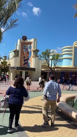 The video shows the entrance area of Warner Bros. Park Madrid on a sunny day. The area is filled with people walking around, presumably heading towards different attractions within the park. There are architectural elements mimicking Hollywood and Beverly Hills. A fountain adds to the scenery. The architecture includes features such as the “Beverly Hills Bakery” building, and the “Alquiler de Carritos y Cajas de Seguridad” building. There is a white arched entrance with the Warner Bros logo on it.