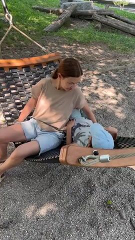 The video shows a woman and a young child relaxing in a hammock outdoors. The woman is wearing a tan t-shirt and denim shorts, while the child is wearing a grey tie-dye shirt and a light blue baseball cap. They are lying in the hammock, which is set up on a gravel surface beneath trees. The scene appears to be a park or a backyard, with a playground-like structure made of logs in the background. Sunlight filters through the trees, suggesting it is daytime. The overall mood seems peaceful and relaxed. The woman is looking down at the child, who is lying on their side. No text is visible in the video. The location is identified as Sala Capriasca, Switzerland.