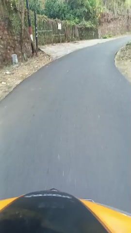 A first-person perspective video shows a road winding through a rural area. The road surface is dark and wet, suggesting recent rain. On the left side of the road, there is a retaining wall made of stone and earth, topped with vegetation. A wire fence is visible along the top of the wall. On the right side, the terrain is covered with dry leaves and sparse vegetation, with some banana trees further in the distance. The sky is bright, indicating daylight. The camera appears to be mounted on a moving object, likely a vehicle or bicycle, given the motion blur. There are no people or animals visible in the frame.