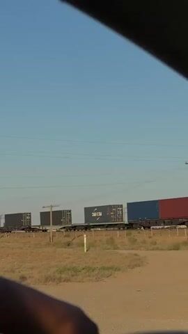 A freight train carrying shipping containers moves across a dry, grassy landscape under a clear blue sky. The train consists of multiple cars, each loaded with one or two large, rectangular containers. Some containers are dark gray, while others are blue and red. One container prominently displays the text "CMA CGM," a shipping company.

The scene appears to be set in a rural or semi-arid location, with sparse vegetation and a dirt road in the foreground. Utility poles and power lines run parallel to the train tracks, suggesting a developed transportation route. The lighting indicates it is late afternoon or early evening, with the sun casting a warm glow on the landscape. The overall impression is one of movement and long-distance transport in a vast, open environment.