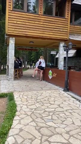 The video captures an outdoor scene at what appears to be a restaurant or commercial area, featuring a wooden building with stone pillars and a paved pathway made of irregular stone slabs. The main subject is a young woman with long blonde hair, dressed in a white sweatshirt, black shorts, and black sandals. She walks briefly and then pauses to take a picture with her smartphone, pointing it towards the right side of the frame. Another woman in a white and blue top is visible behind her. Further in the background, several other individuals are seated or standing near a wooden bar-like structure.

Above the pathway, a wooden sign hangs, reading "Vine and souvenir shop," indicating retail services are available. On a brown wooden fence to the right, a larger wooden sign features a mountain motif and the text "NACIONALNI RESTORAN CRNO JEZERO," which translates to "National Restaurant Black Lake." The name "Nikšićko" is also partially visible on the fence, likely an advertisement for a local beer. The setting is outdoors with trees visible in the background, suggesting a natural or park environment. The lighting indicates it is daytime, and the weather seems mild and pleasant. The overall activity suggests a relaxed atmosphere, with people exploring and enjoying the location. The location context of Pitomine, Montenegro, aligns with the "Black Lake" sign, a well-known site in Durmitor National Park.