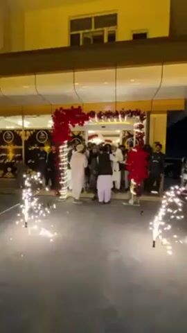 The video depicts a celebratory gathering of men at the entrance of a modern building in Kandahar, Afghanistan, at night. The building has a light-colored facade with large windows on the upper floor, and its entrance is brightly lit. The entrance is elaborately decorated with red floral arrangements or foliage and white string lights, forming an archway.

A group of men, mostly dressed in traditional shalwar kameez, with some wearing vests and turbans, are congregated directly in front of the entrance. They appear to be greeting each other and interacting. On the ground in front of the gathering, several sparklers are actively emitting bright sparks, adding to the festive atmosphere. A white SUV is partially visible parked on the right side of the frame. To the left of the entrance, a sign with visible Arabic script, possibly reading "مدرسه صالون" (Madrasa Salon) or similar, is illuminated, suggesting the opening or celebration of a new establishment. The overall scene conveys a sense of festivity and community engagement.