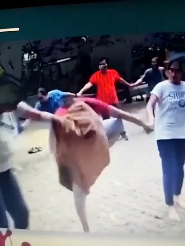 A group of people are holding hands in a circle, some are wearing masks. The people are engaged in some sort of activity or game that involves a large piece of fabric. One person in the center appears to be attempting to wrap the fabric around themselves. The environment appears to be outdoors with some trees and a paved ground.