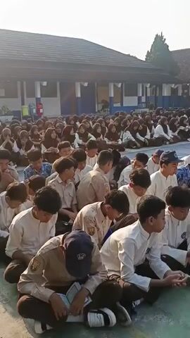 A large group of students, predominantly male, are sitting cross-legged on a paved outdoor surface, likely a schoolyard or sports court. They are wearing uniforms, primarily white shirts and brown pants or skirts, with some wearing hijabs. The students appear to be engaged in reading from books or papers, with many looking down at their laps. The setting is a school with buildings visible in the background, characterized by brick and blue painted walls and a dark roof. The sun is shining, casting shadows and indicating it is daytime. Some students in the foreground are more clearly visible, some wearing caps. The overall atmosphere suggests a communal activity such as a prayer, recitation, or study session.