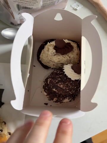 The video shows a small white box containing two fancy cupcakes, one covered in white chocolate shavings and the other in dark chocolate. The box is open, and a hand can be seen holding the opening flaps. To the left of the box is a paper bag with a logo and a half-eaten chocolate chip scone inside. There's also a spoon, a wooden utensil, and packaging in the background, all on a white surface.