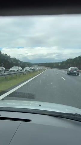 The video shows a view from inside a car driving on a highway. The highway has multiple lanes and is lined with trees on either side. There is moderate traffic on the highway, with cars visible in the same direction of travel and in the opposite direction. The sky is cloudy. There are no people visible in the video.