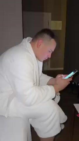 A man in a white terry cloth bathrobe is sitting on the edge of a bed, looking at his phone. He turns his head toward the camera and looks at it briefly. There is a small table to his right, with some items on it. There is a closed door behind him, and the room is dimly lit.