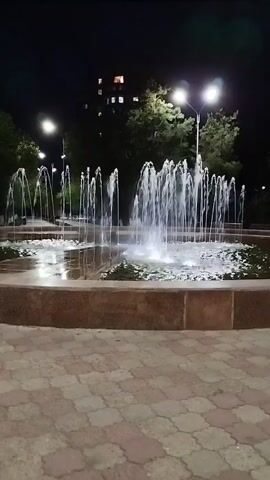 THE VIDEO SHOWS A FOUNTAIN AT NIGHT. THE FOUNTAIN IS IN A POND WITH MARBLE EDGING. THE GROUND AROUND THE FOUNTAIN IS PAVED WITH RED AND TAN TILES. THERE ARE STREETLIGHTS AND TREES IN THE BACKGROUND, AS WELL AS A BUILDING WITH LIGHTS ON IN SOME WINDOWS. THE FOUNTAIN HAS MULTIPLE JETS OF WATER SHOOTING UPWARDS, ILLUMINATED BY LIGHTS BENEATH THE WATER.