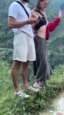 A man and a woman stand side-by-side on a grassy bank beside a shallow body of water, possibly an irrigation ditch, set against a backdrop of lush green mountains in Nậm Dịch Commune, Vietnam. The man, on the left, wears a white t-shirt, beige shorts, and sandals, and is looking at his phone. The woman, on the right, wears a black crop top, grey wide-leg pants, and sandals, with a red jacket draped over her shoulder. She is also holding a phone and gesturing with her left hand while looking slightly away. The scene is brightly lit, suggesting daytime with natural light. They appear relaxed and engaged in conversation, as indicated by their postures and the audio.