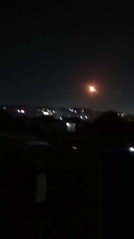 The video, shot at night, shows a cityscape in the distance with a few lights from buildings and a brighter street lamp. The foreground is dark, obscuring specific details, though shapes suggest trees or bushes. The camera is handheld and shaky, as evidenced by the unstable video frame. There are no discernible people or interactions visible. Overall, the scene is dimly lit and out of focus, presenting a night cityscape with dominant darkness in the foreground.