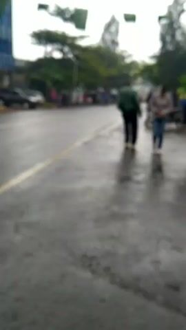 Here is a description of the video:

The video shows a street scene on a rainy day. The road is wet and shiny, reflecting the light. A yellow line marks the edge of the road. Several people are walking along the side of the road, and vehicles, including a van and motorcycles, are driving by. Some people are using umbrellas. A building is visible in the background, and green banners are strung across the street. Overall, the scene depicts a typical rainy day in a busy urban environment.