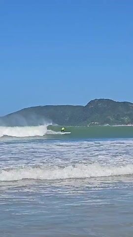 Here's a description of the video:

The video shows a surfer riding a wave on a sunny day. The surfer is wearing a yellow vest and is standing on a white surfboard. The wave is breaking behind the surfer, and there is a lot of foam and spray in the air. In the background, there is a green, hilly landscape under a clear, blue sky. The foreground shows the choppy water closer to the beach.