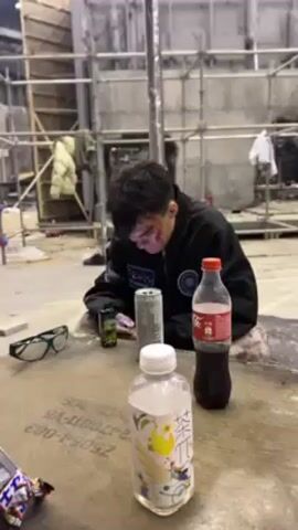 The video shows a person sitting at a table in a messy, industrial-looking environment. The person has fake blood on their face. The table has various items on it, including drinks, snacks, glasses, and a book. The person smiles at the camera, then holds up a smartphone displaying a photo of a man with tattoos and a white robe. The photo has a caption: "POV: You lock in after seeing Maki's take on the Zenin clan." Other people can be seen in the background walking around.