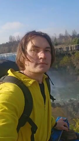 A person wearing a yellow jacket and a backpack is standing in front of a waterfall, taking a selfie. The waterfall is wide and powerful, with mist rising from the base. The person appears to be on a cliff or raised area overlooking the waterfall. There are trees and foliage in the background. It is a sunny day.