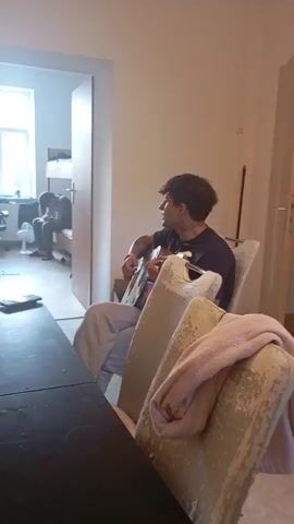 A young man with dark curly hair sits in a chair playing an acoustic guitar. He is wearing a dark t-shirt and light-colored pants. In the background, another person is sitting on a bunk bed in what appears to be a room with a window and a fan. The foreground shows a table and two chairs with a blanket draped over one of them. The lighting is dim and the scene appears to be indoors.