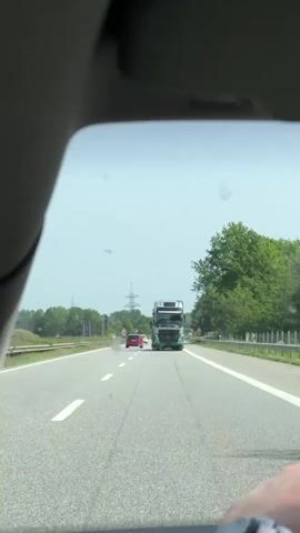 The video shows a view through the windshield of a car driving on a highway. In the distance, a large semi-truck is travelling in the same direction, along with a red car further ahead. The sky is clear and there are trees lining the side of the road. The road has white lane markers. An electrical power pole is visible in the distance. The car is travelling on the right lane. The video appears to be taken during daylight hours.