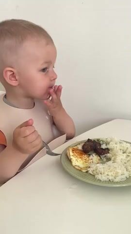 A toddler wearing an orange shirt and a beige bib is sitting at a white table, eating rice, a piece of fried egg, and some meat from a green plate with a fork and their hands. The toddler is looking down at the food. The background is a plain white wall.