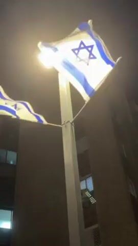 The video shows two Israeli flags waving in the wind at night, illuminated by a street lamp. The flags are attached to a pole in front of a tall building with some visible windows. The sky is dark.