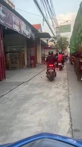 The video shows a perspective from a rider on a blue motorcycle. In front, three motorcycles are traveling down a paved road. The lead motorcycle is black and has a rider wearing a black helmet and a red shirt. The second motorcycle is red with a rider wearing a black helmet and a white shirt. The third motorcycle is black and is partially obscured by the second. To the right of the road, a large green truck is parked. Further to the right, there are trees and utility poles. On the left side of the road, there are buildings with awnings and signs, and some store displays. One sign on the left advertises "Anik". A street sign with directional arrows indicating "Blora, Cepu", "Kuwu, Doplang", and "Karang Asem" is visible above the trucks. The sky is visible and appears to be dusk.