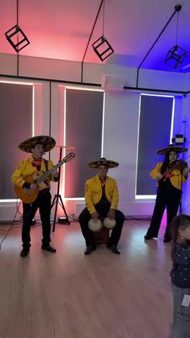 A mariachi band is performing in a room with a wooden floor. A man in a yellow jacket, black pants, and a sombrero is playing a guitar and singing. To his right, another man in similar attire is sitting on a box and playing bongos. Further to the right, a woman in a yellow jacket and sombrero is playing a violin. The room has modern lighting, with vertical strips of red and blue light on the back wall. There are speakers mounted on the wall and cube-shaped light fixtures hanging from the ceiling.