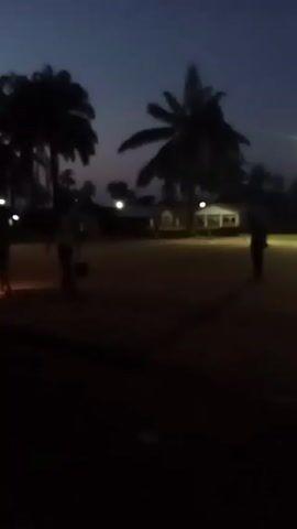 The video shows a dark outdoor scene at twilight. There are several silhouetted figures walking across a sandy or grassy area. Palm trees are visible in the background against the darkening sky. There is a building with lights on in the background. The video is shot from under a roof, looking out from a shaded area.