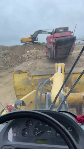 The video provides a first-person view from the cab of a yellow wheel loader at a construction or quarry site in Hristiyanovo, Bulgaria. The driver's seat, steering wheel, dashboard, and the front bucket of the loader are visible, along with hydraulic cylinders.

In the background, a large pile of broken rocks and earth dominates the scene under an overcast sky. On top of this pile, a yellow Volvo excavator is positioned next to a large red mobile rock crusher or screener, suggesting an active material processing operation. Another smaller red excavator is visible further in the distance on the same elevated terrain.

The video briefly pans to the left, showing more of the dusty, rugged landscape and a yellow camera-like device mounted on the side of the loader. It then returns to the frontal view of the excavator and crusher. The final shot is a reflection in the side mirror, revealing an expansive, barren area of the site with some distant structures, possibly temporary buildings or additional equipment. The ground is rough and dusty, characteristic of a heavy industrial worksite. No people are visible, only the heavy machinery at work or in standby.