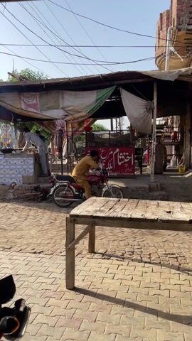 The video shows a street scene in what appears to be a South Asian country. The street is paved with brick and lined with buildings on both sides. Underneath a simple shelter structure, two men are working, one standing near a cooking area with a large pan, possibly frying food, and the other sits on a red motorcycle parked in front of the structure. A third man walks past them, pushing a cart down the street. Several motorcycles are visible, parked or moving in the background. A sign with a menu is displayed near the street. The scene is recorded from a slightly elevated perspective, possibly under another shelter or canopy, with a partial view of a motorcycle handlebar and mirror in the foreground. The sky is clear and sunny.