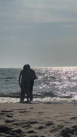 Two figures stand on a sandy beach at the edge of the water, facing the ocean. Sunlight glints off the water's surface, creating a sparkling effect. The figures appear to be interacting, with one seemingly leaning on or embracing the other. Waves gently lap at their feet as they stand near the waterline. The sky is partially cloudy, with the sun visible behind the clouds. The scene is silhouetted against the bright sky and water.