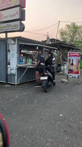 The video shows an outdoor scene at dusk with two people standing at a small roadside kiosk. A man in dark shorts and a t-shirt is facing the kiosk, and a woman in a dark top and pants is standing behind him, leaning against a motorcycle. The kiosk appears to be a temporary structure made from a shipping container, with a counter and shelves displaying bottles and other items. A sign above the kiosk reads "SURGA UMUM 13 RIBU" and "lumer hotpan COKLAT manisnya kamu". To the right of the kiosk, there is another sign with a picture of grapes and text that says "ES ASSU ANGGUR". The ground is unpaved and appears to be dirt or gravel. Trees are visible in the background. The lighting suggests it is either early morning or late evening.