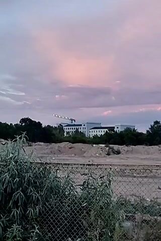 This media file captures a wide shot of a construction site under a dramatic, colorful sky, likely during sunset. The foreground features a chain-link fence with some vegetation growing around its base. Beyond the fence lies a vast, uneven landscape of dirt and debris, suggesting ongoing or recently completed demolition or excavation.

In the mid-ground, a large, white, multi-story building stands prominently, appearing to be an office building or a similar institutional structure. To the left of the building, a construction crane is visible, its arm extended and illuminated by several white lights, indicating work may have continued into the evening or is prepared for early morning. The building is surrounded by a dense line of dark green trees.

The sky is a dominant feature, painted with soft hues of pink, purple, and pale blue, indicative of twilight. Wispy clouds are scattered across the sky, some catching the last rays of sunlight and glowing with a vibrant pink. The overall mood is one of transition and potential, with the juxtaposition of the raw construction site and the serene, beautiful sky. There are no visible people or text in the media file. The location context of Dolinka, Kyrgyzstan, suggests a potentially arid or semi-arid environment, which could be consistent with the dusty landscape.