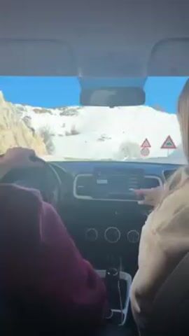 From the perspective of inside a car, a person wearing a maroon sweatshirt is driving on a winding mountain road covered in snow. A passenger is seated to the right. The weather is sunny and clear. The car has a dashboard screen and visible climate control buttons.  A road sign warning of curves is visible outside the front windshield.  The car approaches a short tunnel carved into the snowy mountainside.