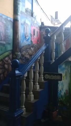 The video shows a small courtyard. The stairs leading up on the left of the frame have a dark blue railing and painted white balusters. The stairs are painted a lighter shade of blue, and the walls nearby are painted with murals. There is a sign that says "La Guachafita, Bakery Burger & Bar." There is a support column to the right of the stairs that also has a mural painted on it. To the right of the column, there is a green wall with a window that has bars in it. Inside the window, there are bottles and other objects displayed. To the right of the green building, there is a gate leading to a courtyard with yellow and blue walls. There is a sign visible through the gate.