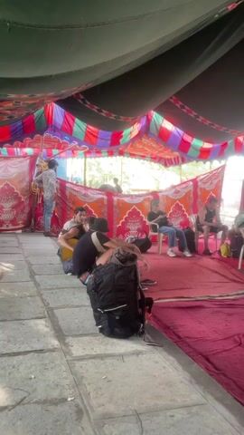 The video shows a group of people sitting and standing under a tent-like structure. The structure is decorated with colorful fabric and has a red carpet on the ground. Some people are sitting in chairs, while others are sitting on the floor. Two men walk towards the camera, one in a tank top and blue shorts, and the other in a white shirt and black shorts. There are backpacks and other personal items scattered around the area. A man can be seen adjusting the tent's ropes in the background. The scene gives the impression of a casual gathering or waiting area.