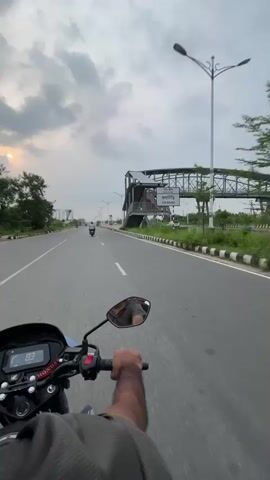 A person rides a blue Honda motorcycle on a gray paved highway. The speedometer reads 82-84 kilometers per hour. The rider’s right arm and hand grip the throttle. Other vehicles are visible on the road ahead, including another motorcycle and a small green three-wheeled vehicle. The sky is cloudy, and the sun is visible through a gap in the clouds. On the right side of the road, there are white streetlights, a black and white striped barrier, and a metal pedestrian overpass. A sign next to the overpass has the word “JOLS” visible. The scenery to the left includes trees and a partially visible billboard in the distance.