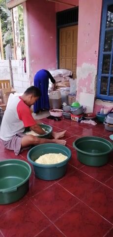 A young boy, wearing a red shirt and striped shorts, sits on a red tiled floor and mixes a pale yellow substance in a green basin with his hands. He is facing to the right and looking down at his task. To his right, another green basin contains a larger amount of the same yellow substance. Further to the right, a third green basin is empty. In the background, to the left, a woman wearing a blue head covering and a long blue dress stands and appears to be handling large white bags. She is facing away from the camera and towards a wall. Behind her are various containers and bags. The environment appears to be an outdoor or semi-outdoor covered area, with a pink-painted wall and a window with blue trim visible. The floor is made of red tiles.