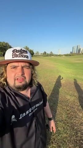 The video shows a man standing on a grassy field in front of what appears to be a large park area, and in the background, a cityscape. The man has long curly hair, a beard, and wears a hat and a black jacket with white lettering. He is holding the camera and speaking, pointing upwards towards a large balloon in the sky. The balloon is spherical and features the words "LIVE MORE" and Hebrew text, possibly related to investments. The setting appears to be a sunny day with a clear blue sky.