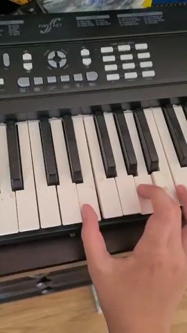 A person's hand plays a keyboard, pressing keys to produce a melody. The keyboard is black and white with some visible wear on the white keys. The background is blurry but appears to show a desk or shelf with various items.
