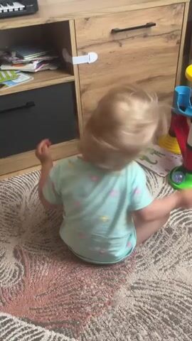 A toddler with blonde hair and wearing a light blue t-shirt with pastel colored polka dots sits on a rug in front of a wooden cabinet. The toddler is turning their head from left to right, with their body slightly twisted to the side. There is a toy with colorful rings on the floor to the right of the toddler, and the cabinet has several shelves and drawers. The rug has a pattern of pink and beige lines.