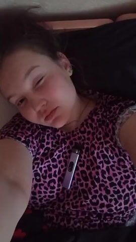 Here is a concise description of the video:

A woman is lying down wearing a pink and black leopard print shirt. There is a silver vape pen on her shirt. She is making a kissing face. The video is taken from a high angle, looking down at her.
