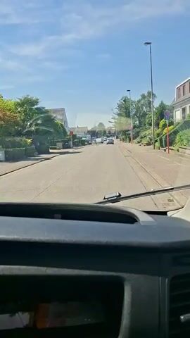 A video recorded from inside a vehicle shows a view of a residential street on a sunny day. The camera is positioned behind the dashboard, with the windshield and side window visible. The road ahead curves to the right, with houses and trees lining both sides. Several cars are visible on the road, some parked and some in motion. The sky is blue with scattered white clouds. The video appears to be a dashcam recording, capturing the journey down the street. The interior of the vehicle is dimly lit in comparison to the bright exterior.