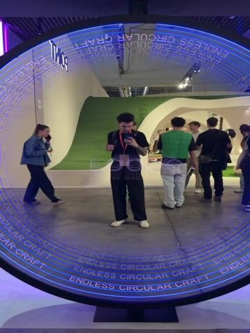 The video captures an indoor exhibition or event in Rho, Italy, featuring modern design and lighting.

Initially, a young man with a beard and tattoos, wearing a black t-shirt and pants with an ID badge, stands before a large, circular, reflective display. He is holding a phone, seemingly recording himself or the surroundings. The display features concentric blue neon rings that repeatedly show the mirrored and forward text "ENDLESS CIRCULAR CRAFT" and "SCROLL". Other attendees are visible in the background and reflections, walking or standing.

The camera then pans to reveal a lounge or seating area within the exhibition. This space is illuminated by vibrant purple and blue neon lights, with a large cloud-shaped light fixture on the ceiling. Several people are gathered in this area: two women sit in bright pink modern chairs, one gesticulating while talking; others are seated on a long, curved blue bench. A small potted tree stands near the seating. People are conversing, relaxing, and one person appears to be on a laptop. The word "loope" is visible on the wall above the blue bench.

The scene depicts a contemporary and engaging atmosphere at what appears to be a design, tech, or art fair, with attendees interacting with the exhibits and each other in a stylish, well-lit environment.