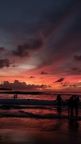 Silhouettes of people stand and wade in the ocean during a vibrant sunset. The sky is a mix of orange, red, and dark grey clouds, reflecting onto the wet sand. Small waves roll onto the beach. Some people are partially submerged in the water. A land mass can be seen in the distance on the horizon. The overall impression is a colorful, serene, and peaceful beach scene at dusk.