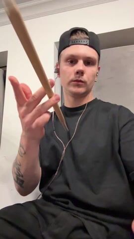 A man in a black t-shirt and backwards black baseball cap is seated and spinning a wooden drumstick in front of his face. He has fair skin and short hair, and he wears wired earphones. A tattoo is visible on his left forearm. The background consists of white walls and a gray panel, suggesting an indoor setting.