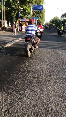 The video shows a street scene in Bligo, Indonesia. The primary subjects are people riding motorcycles and walking along the sidewalk. There are also food stalls with umbrellas and signs visible. The scene is set on a paved road with a sidewalk.

The activity taking place is normal street life. People are on their way somewhere, possibly commuting or running errands. Some are on motorcycles, while others are walking. There appears to be some commercial activity with the food stalls.

Notable details include the time of day, which appears to be late afternoon or early evening, as indicated by the warm, angled sunlight and long shadows. The weather seems pleasant, with no signs of rain. The location cues, such as the style of the stalls and the language on the signs, point to Indonesia. The mood seems relaxed and ordinary.

Visible text includes "LAJUR PRIORITAS BUS TRANS JATIM JAM OPERASIONAL" on a blue sign above the road, indicating a priority bus lane for Trans Jatim. There is also a pink sign with "TAHU PETIS" and "ARRAY" which likely advertises a local food item. The license plate of the motorcycle in the foreground reads "M 3099 ZT 10-23".