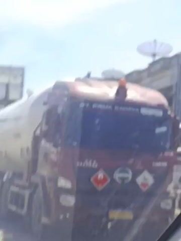 The video shows a street scene with various trucks. A large white tanker truck with the inscription "PERTAMINA" is in the foreground. A red truck with a container is behind it. The camera then pans to show a white truck with a blue frame and an Isuzu logo. The scene is bright and sunny. The trucks appear to be driving on a road in a town or city.