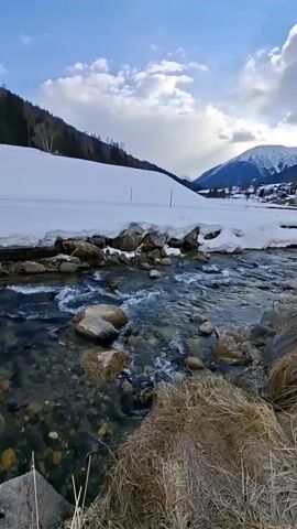 The video shows a picturesque winter scene with a clear, shallow stream flowing through a snowy landscape. The water rushes around and over rocks, and the stream is flanked by dry grass and patches of snow. A large snow-covered field stretches out on one side of the stream, leading up to a forested hill. In the distance, a snow-capped mountain looms over a small village, with a blue sky dotted with clouds visible above. The scene is tranquil, showcasing the contrast between the flowing water and the serene, snow-covered surroundings.